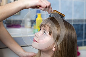 Mother hand with brush combing long fair hair of cute child girl after bath on blurred interior background