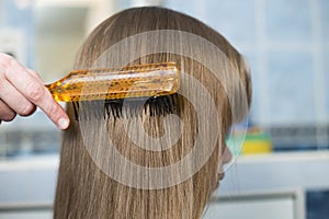 Mother hand with brush combing long fair hair of cute child girl after bath on blurred interior background