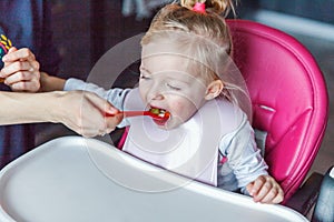 Mother hand feeding infant girl with spoon in high baby chair in kitchen at home