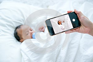 Mother hand hold mobile phone to take a photo of newborn baby during she is sleeping on white bed