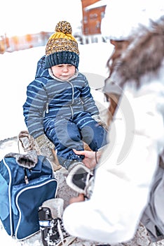 Mother helps woman, in winter, put on shoes skates, son little boy 3-5 years old, train and learn to skate, warm clothes