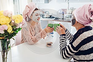 Mother and her adult daughter drinking tea with facial masks applied. Women chilling and talking on kitchen