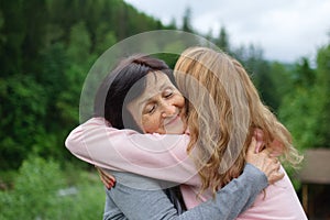 Mother and her adult daughter are hugging spending time together outdoors over landscape of forest and mountains