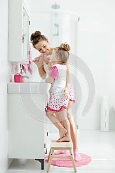 Mother and her daughter brush their teeth with toothbrushes in the bathroom at home. Mom and baby girl in home clothes.