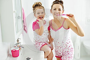 Mother and her daughter brush their teeth with toothbrushes in the bathroom at home. Mom and baby girl in home clothes.