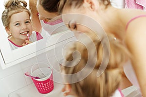 Mother and her daughter brush their teeth with toothbrushes in the bathroom at home. Mom and baby girl in home clothes.