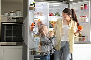 Mother and her daughter with fruits near open refrigerator in kitchen