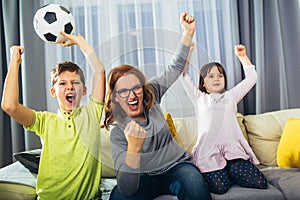 Mother and her kids raising arms and screaming while watching football match and celebrating goal