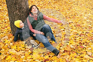 Mother and her little child boy sitting near huge maple tree in autumn park and looking on falling leaves. Fall nature banner with