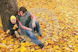 Mother and her little child boy sitting near huge maple tree in autumn park and looking on falling leaves. Fall nature banner with