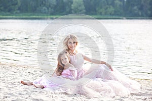 Mother and her little girl enjoying lake view and relaxing on the beach on a sunny day in beautiful dresses. Family lifestyle and