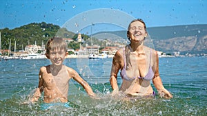 Mother and her son playing and splashing water at the sea beach in slow motion. Family holiday, vacation memories, and