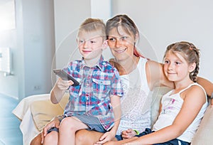 Mother and her sons are watching tv while sitting on a couch at home.Happy mom and her sons on back sofa with TV remote control