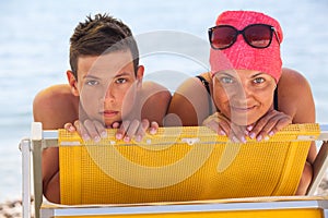 Mother and her teenage son lying on yellow chaise-longue on sea coast. Looking at camera and smiling together