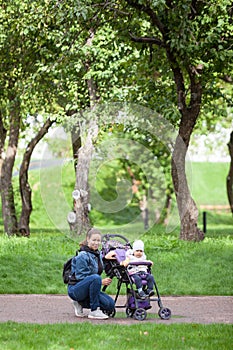 Mother and her toddler daughter walking in garden with a children pram, woman sitting close to girl, vertical image