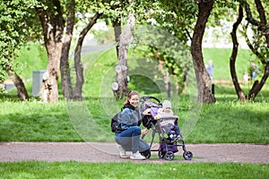 Mother and her toddler daughter walking in garden with infant stroller, woman sitting close to girl, horizontal image