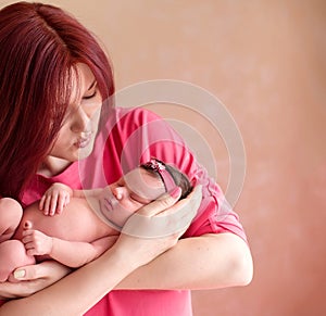 Mother holding in arms newborn daughter in pink hair band and leaning to kiss her baby. Mom and infant child together.