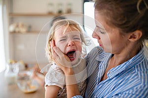 A mother holding a crying toddler daughter indoors in kitchen. photo