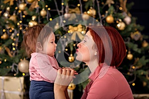 Mother holding hands and kissing infant baby daughter in front of decorated Christmas New Year fir tree. Family winter holidays