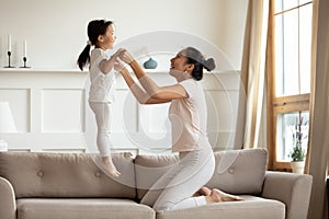 Mother holding hands little daughter while she jumping on couch