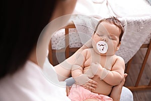 Mother holding her cute little baby with pacifier at home, closeup