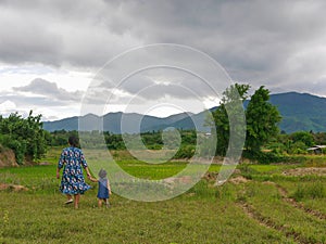 Mother holding her little baby girl`s hand walking in a field in a countryside, watching rainy clouds forming in the sky