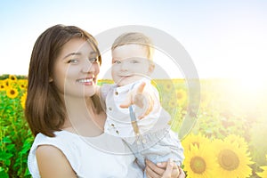 Mother holding her son in arms in sunflowers field. The boy is pointing his finger at the camera and smiling