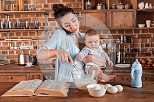 Mother holding her son, talking on smartphone and mixing a dough at the kitchen. family life and multitasking concept photo