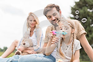 mother holding teddy bear and looking at father and daughter blowing soap bubbles