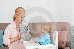 Mother holding teddy bear near cute toddler son in feeding chir
