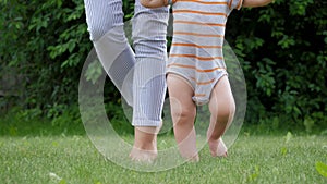 Mother Holds Hands With Her Baby Helping Him Take The First Steps On Green Lawn