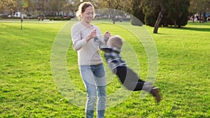 Mother holds her son by arms and whirls with him in park at sunset