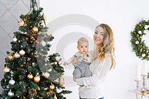 Mother holds her young son in her arms against the background of a Christmas tree in the living room. They are going to celebrate