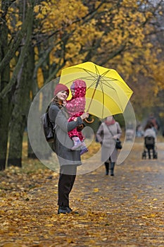 Mother holds a small child in her arms while standing under a yellow umbrella in an autumn park against a background of yellow