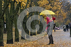 Mother holds a small child in her arms while standing under a yellow umbrella in an autumn park against a background of yellow