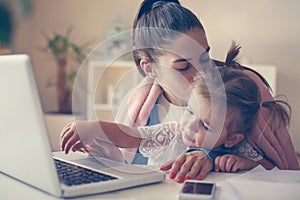 Mother at home kissing her daughter and using laptop toget