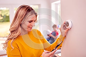 Mother At Home With Son Adjusting Smart Central Heating Thermostat Control With App On Mobile Phone