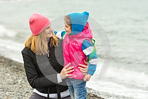 Mother hugging little daughter and tenderly looking at her on beach in cold weather