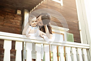 A mother hugs and kisses a little innocent baby girl on the outdoor veranda of an old wooden house.