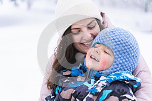 Mother hugs little smiling laughing cute boy kid.Red blush on cheeks.Happy family walking in beautiful winter forest park covered