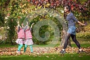 Mother and identical twins having fun with autumn leaves in the park, blond cute curly girls, happy kids, girls in pink jacket