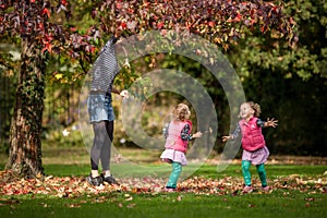 Mother and identical twins having fun with autumn leaves in the park, blond cute curly girls, happy kids, girls in pink jacket