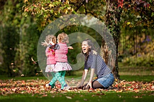 Mother and identical twins having fun with autumn leaves in the park, blond cute curly girls, happy kids, girls in pink jacket