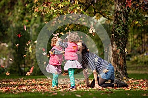 Mother and identical twins having fun with autumn leaves in the park, blond cute curly girls, happy kids, girls in pink jacket