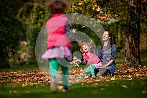 Mother and identical twins having fun with autumn leaves in the park, blond cute curly girls, happy kids, girls in pink jacket