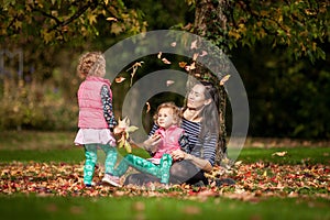 Mother and identical twins having fun with autumn leaves in the park, blond cute curly girls, happy kids, girls in pink jacket