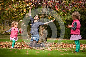 Mother and identical twins having fun with autumn leaves in the park, blond cute curly girls, happy kids, girls in pink jacket