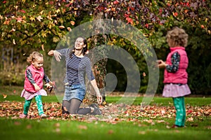 Mother and identical twins having fun with autumn leaves under the tree in the park, blond cute curly girls, happy family