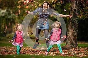 Mother and identical twins having fun with autumn leaves under the tree in the park, blond cute curly girls, happy family