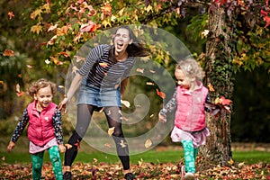 Mother and identical twins having fun with autumn leaves under the tree in the park, blond cute curly girls, happy family